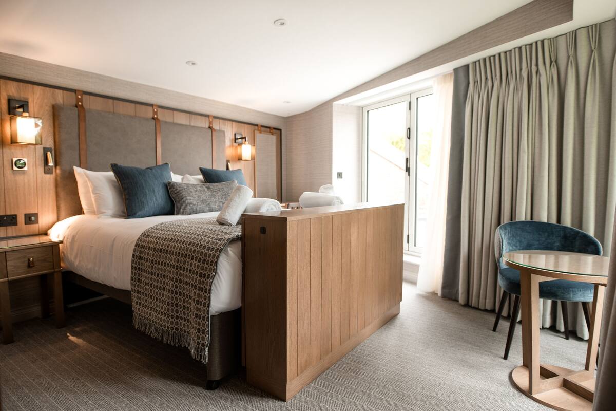 A modern hotel room with a wooden headboard, a large bed with multiple pillows, a bedside table, a small desk with a chair, and a view of the outdoors through a sliding door.