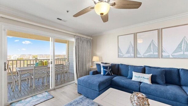 Ein Wohnzimmer mit einem blauen Sofa, einem Deckenventilator und Blick auf einen Balkon mit Gartenmöbeln.