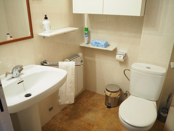 Une salle de bain avec un lavabo, des toilettes et une poubelle. On y trouve des étagères avec des articles de toilette et une serviette accrochée au mur.