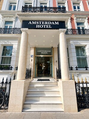 The entrance to the Amsterdam Hotel with a grand facade, large glass doors, and a set of stairs leading up to the entrance. - Amsterdam Hotel (London)