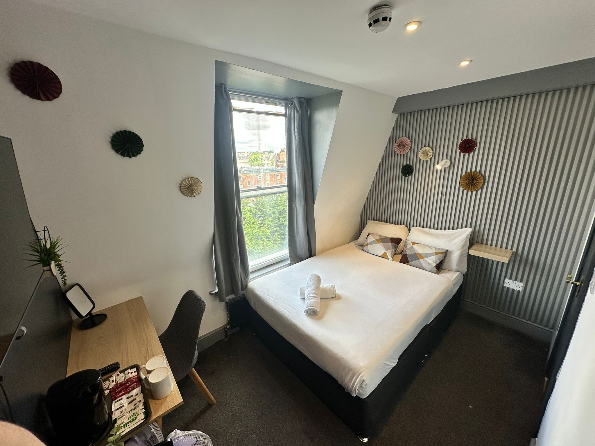 A modern hotel room with a bed, a desk with a chair, a small table with a plant, and a window with curtains.
