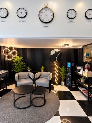A modern lobby with clocks indicating different cities, seating area with two armchairs and a round table, and a coffee machine. - Amsterdam Hotel (London)