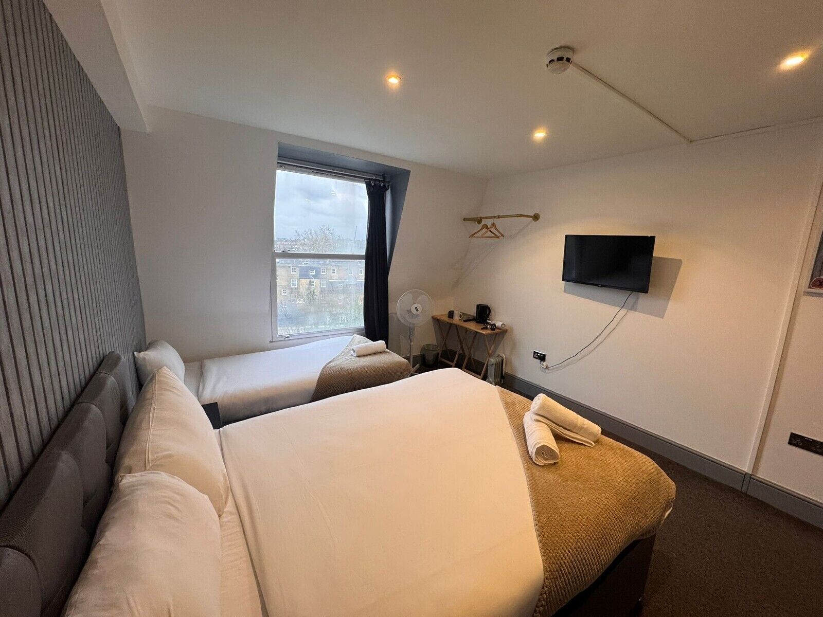 Ein Hotelzimmer mit zwei Betten, einem Fernseher, einem kleinen Tisch mit Ventilator und einem Fenster mit Blick auf Gebäude.
