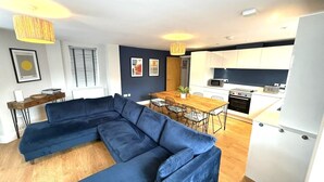 A modern living room with a sectional sofa, a dining table with chairs, a kitchen area with white cabinets and appliances, and a small side table with a lamp. - Bernard Point (Southampton)