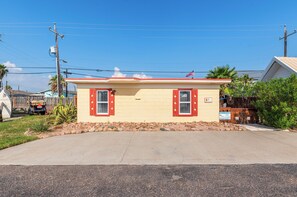 A small, single-story building with a beige exterior and red shutters, located at the end of a driveway.