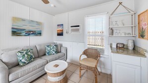 A cozy living room with a grey sofa, wicker chair, and a small table. A painting of a boat on water is on the wall, and there is a window with blinds. - This attractive cottage at Spanish Village in Port Aransas is a free standing ho (Port Aansas)