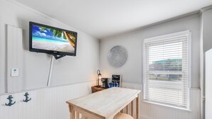 A room with a wooden table, a television mounted on the wall, a round decorative plate, and a window with blinds. - This attractive cottage at Spanish Village in Port Aransas is a free standing ho (Port Aansas)