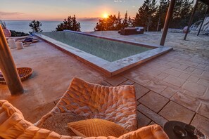 A pool area with a view of the sunset over the sea, surrounded by stone flooring and outdoor furniture.