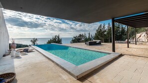 A rectangular pool with clear blue water, surrounded by a patio area with outdoor furniture and a view of the sea.