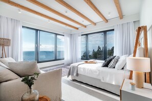 A modern bedroom with large windows, a bed with white linens, a bedside table with a lamp, and a view of the sea.