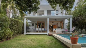 Ein modernes, zweistöckiges Haus mit überdachter Terrasse, Schwimmbad und üppiger Bepflanzung.