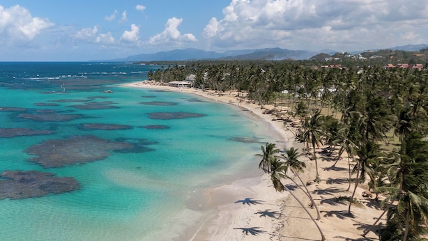 Ein Strand mit klarem türkisblauem Wasser, Palmen und einem fernen Gebirge.