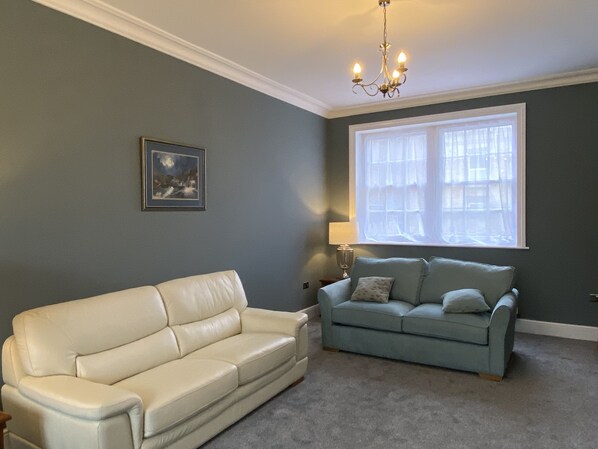 A living room with a beige sofa, a light blue sofa, a chandelier, a window with sheer curtains, and a framed picture on the wall.