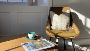 A chair with a patterned pillow, a cup and saucer, and a magazine on a wooden table by a window with a plant. - Red Barns Bed & Breakfast (Redcar)
