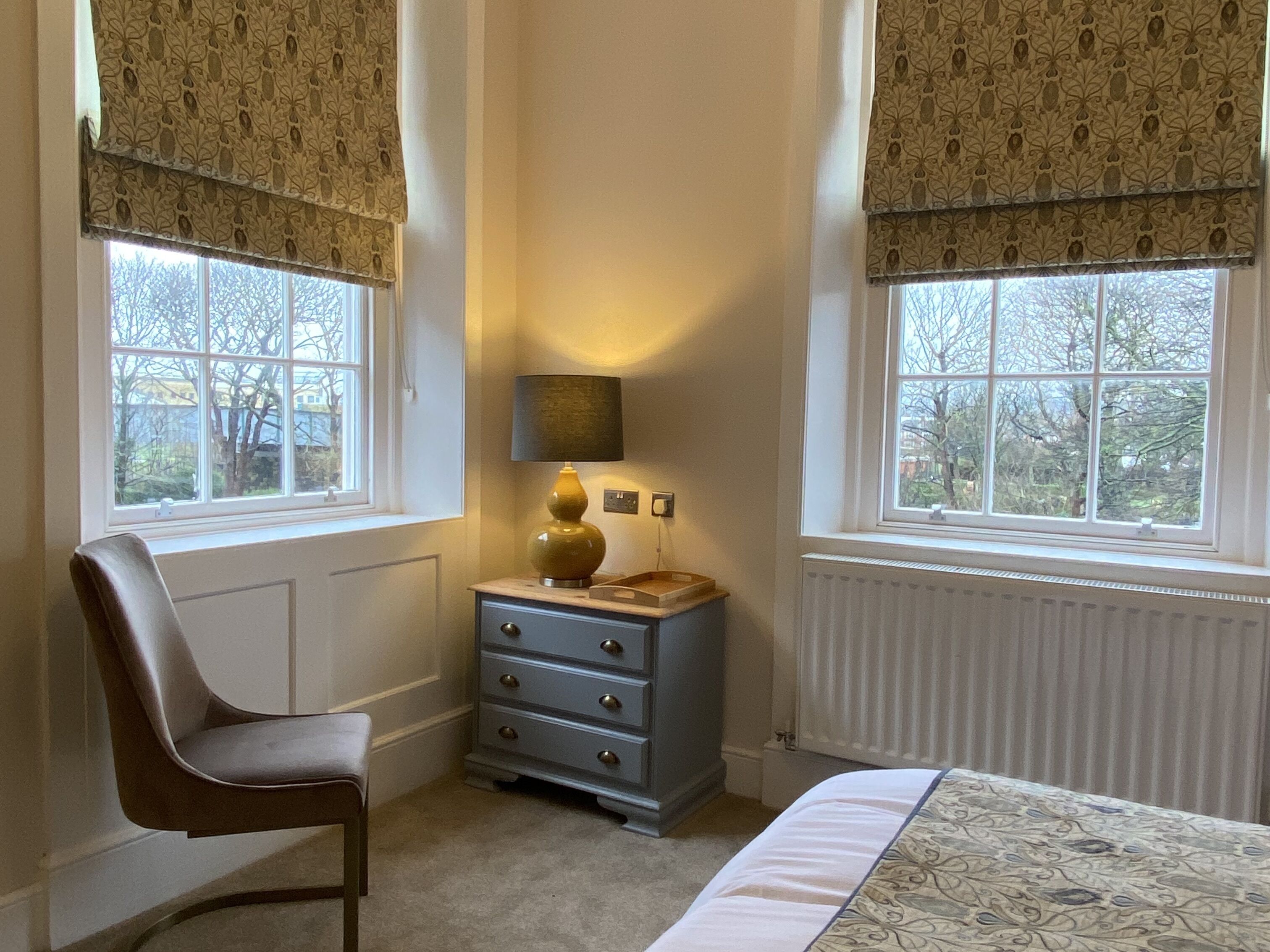 Une chambre à coucher comprenant un lit, une commode, une chaise et deux fenêtres ornées de rideaux à motifs.