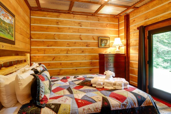 A wooden cabin bedroom with a bed, pillows, a quilt, a nightstand with a lamp, and a window with a view of trees.