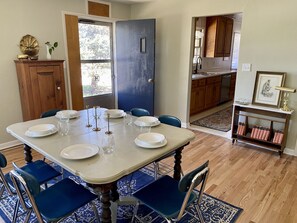 Une salle à manger avec une grande table dressée pour huit personnes, entourée de chaises bleues. On aperçoit un buffet en bois et une porte bleue à l’arrière-plan.