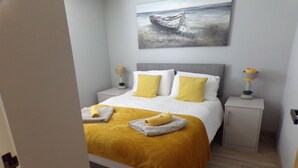Un lit bien fait, avec une couverture et des oreillers jaunes, une table de chevet avec une lampe, et un tableau représentant un bateau accroché au mur.
