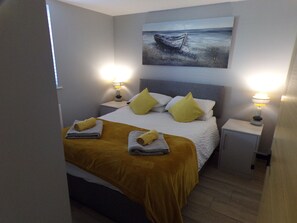 A neatly made bed with a yellow throw blanket and pillows, two bedside lamps, and a painting of a boat on the wall. - 4thedome (New Milton)