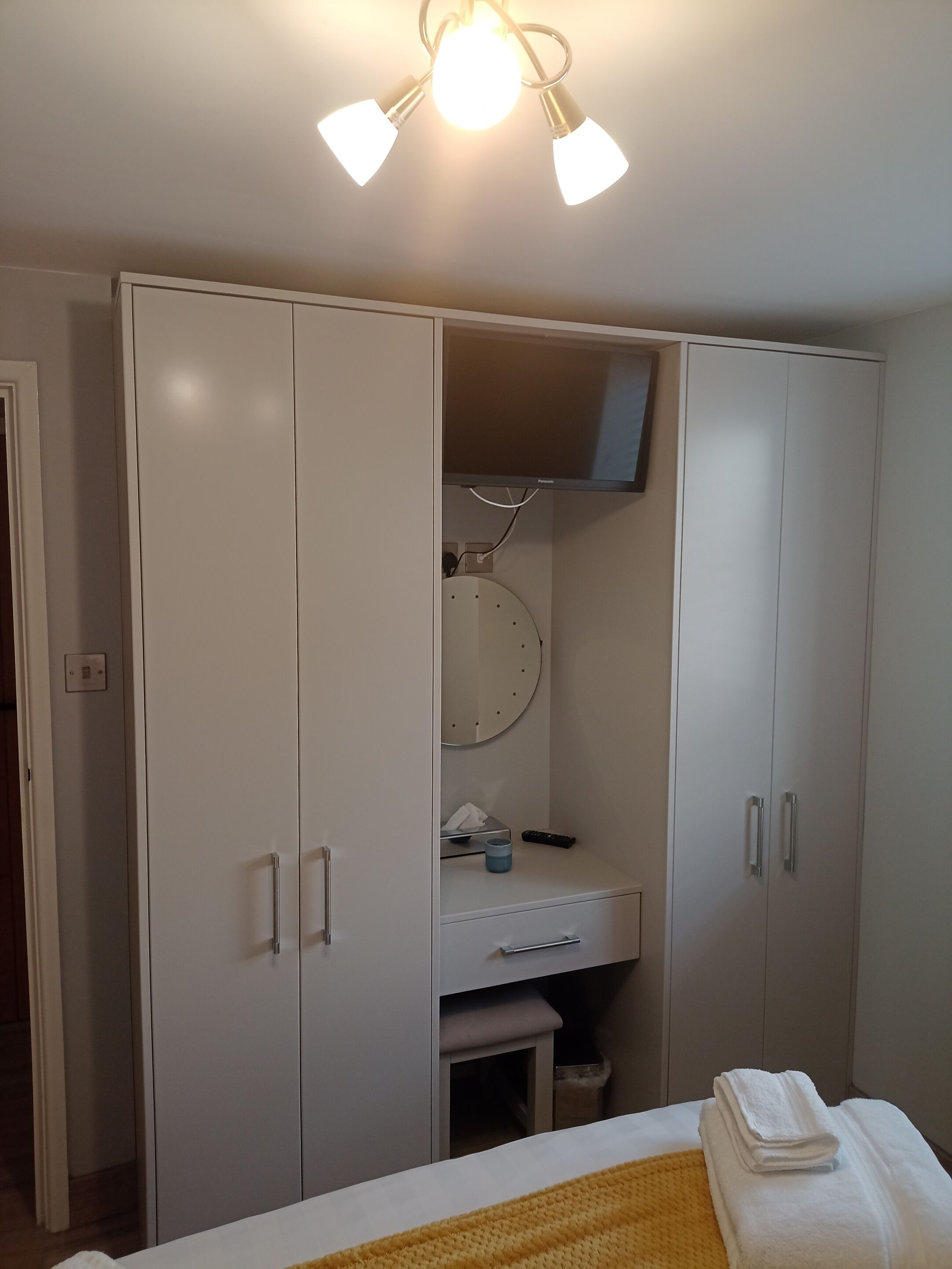 Un dormitorio con cama, armario, escritorio con espejo y lámpara en el techo.