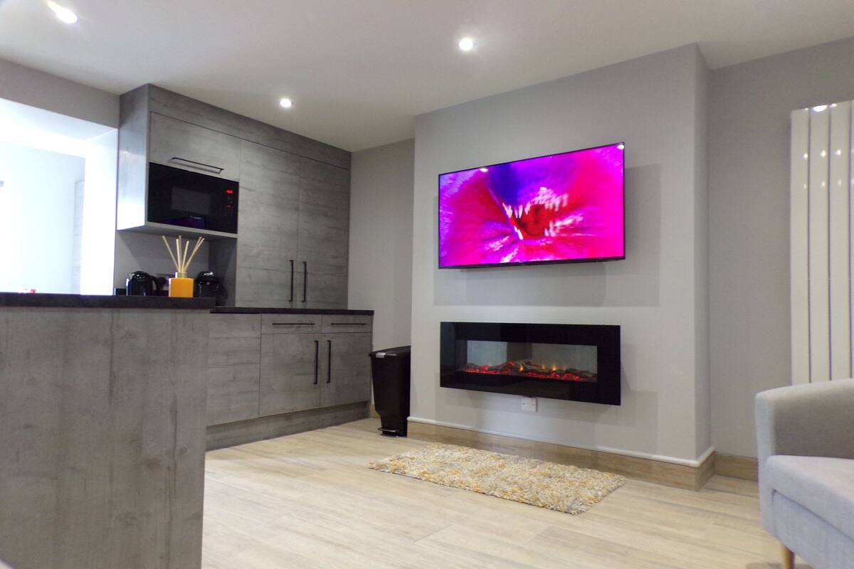 Una cocina moderna con microondas integrado, un televisor de pantalla plana sobre una chimenea y un armario gris con encimera negra.
