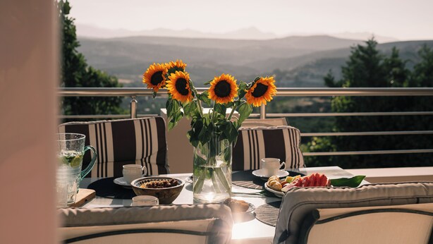 A dining table with a vase of sunflowers, a glass pitcher, and a bowl of food, set against a scenic mountain backdrop. - Blue Sky Villa in Crete (Επάνω Βαθειά)