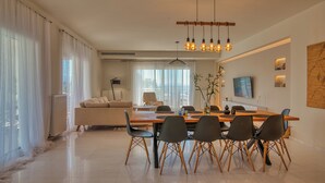 A modern living room with a dining area, featuring a sofa, a dining table with chairs, a TV, and a hanging light fixture. - Blue Sky Villa in Crete (Επάνω Βαθειά)