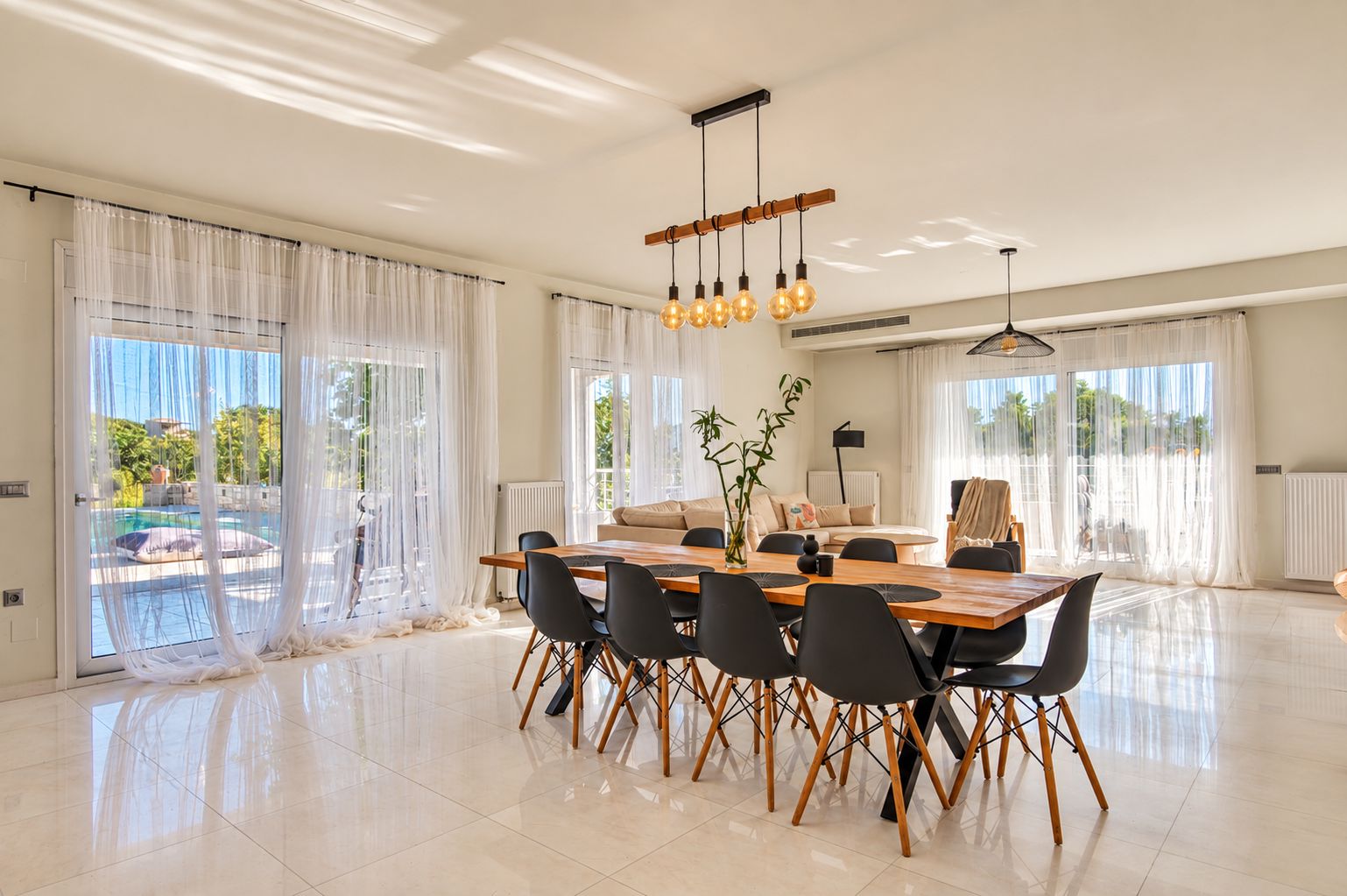 A spacious dining area with a wooden table and chairs, large windows with sheer curtains, and a ceiling light fixture.