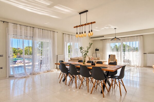 A spacious dining area with a wooden table and chairs, large windows with sheer curtains, and a ceiling light fixture.