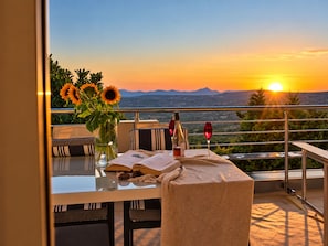 A balcony with a table set for two, a book, a bottle, and a glass of red wine, overlooking a sunset and mountains.
