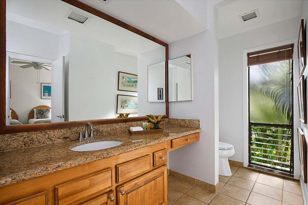 Un baño con encimera de granito, muebles de madera, un espejo grande y un inodoro con puerta corredera de vidrio a un balcón.