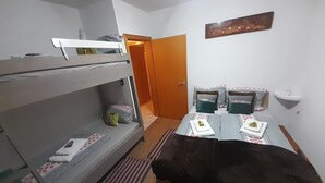 Een studentenkamer met stapelbedden, een klein tafeltje met een plaid, een nachtkastje met een lamp en een deur naar een andere kamer.