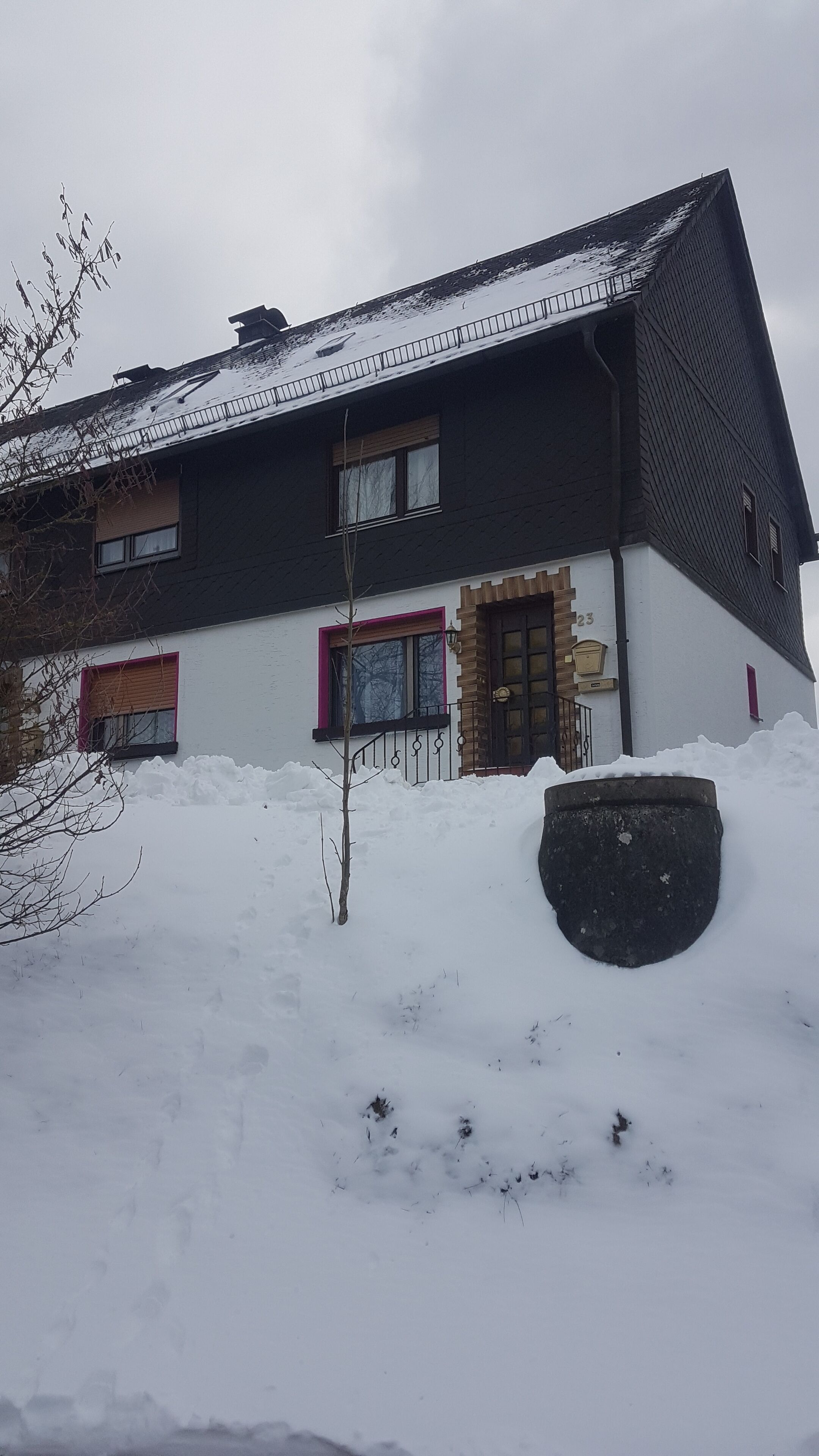 Een huis met twee verdiepingen, een met sneeuw bedekte dak en een grote zwarte prullenbak ervoor.