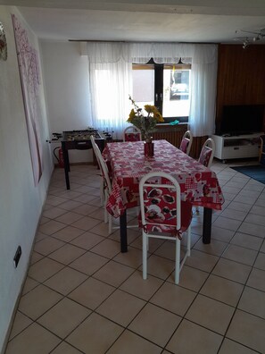 A dining area with a red and white checkered tablecloth, white chairs, a foosball table, and a television on a stand.