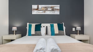 A neatly made bed with white and teal pillows, a beige blanket, and white towels at the foot of the bed. There are two bedside tables with lamps and a painting on the wall above the bed. - Matrix 504 (Cape Town)