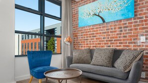 A modern living room with a blue chair, grey sofa, and a round coffee table. A large window offers a view of a brick building. - Matrix 504 (Cape Town)