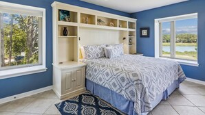 Un dormitorio con cama, estanterías empotradas, ventilador de techo y ventanas amplias con vistas a árboles y un lago.