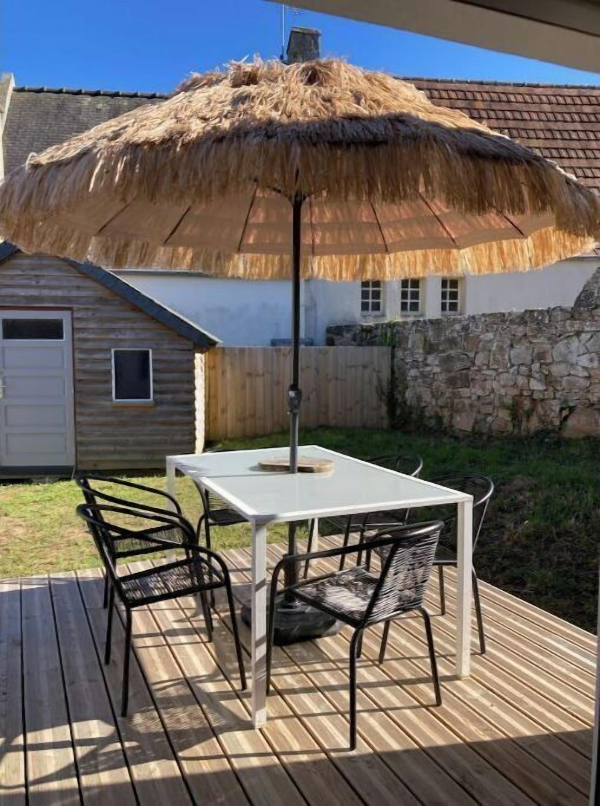 Una terraza de madera con una mesa y sillas, un paraguas de paja y un muro de piedra al fondo.