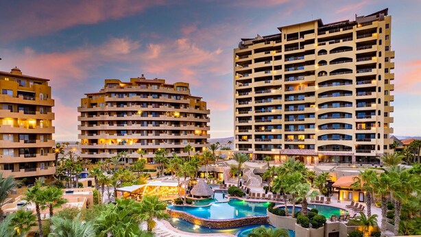 A resort with multiple hotel buildings, a central swimming pool, and surrounding palm trees.