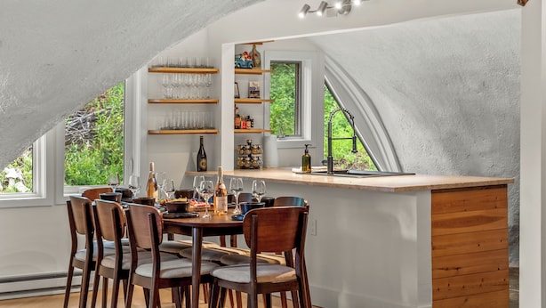 A dining area with a wooden table and chairs, a built-in shelf with glassware, and a view of greenery outside.