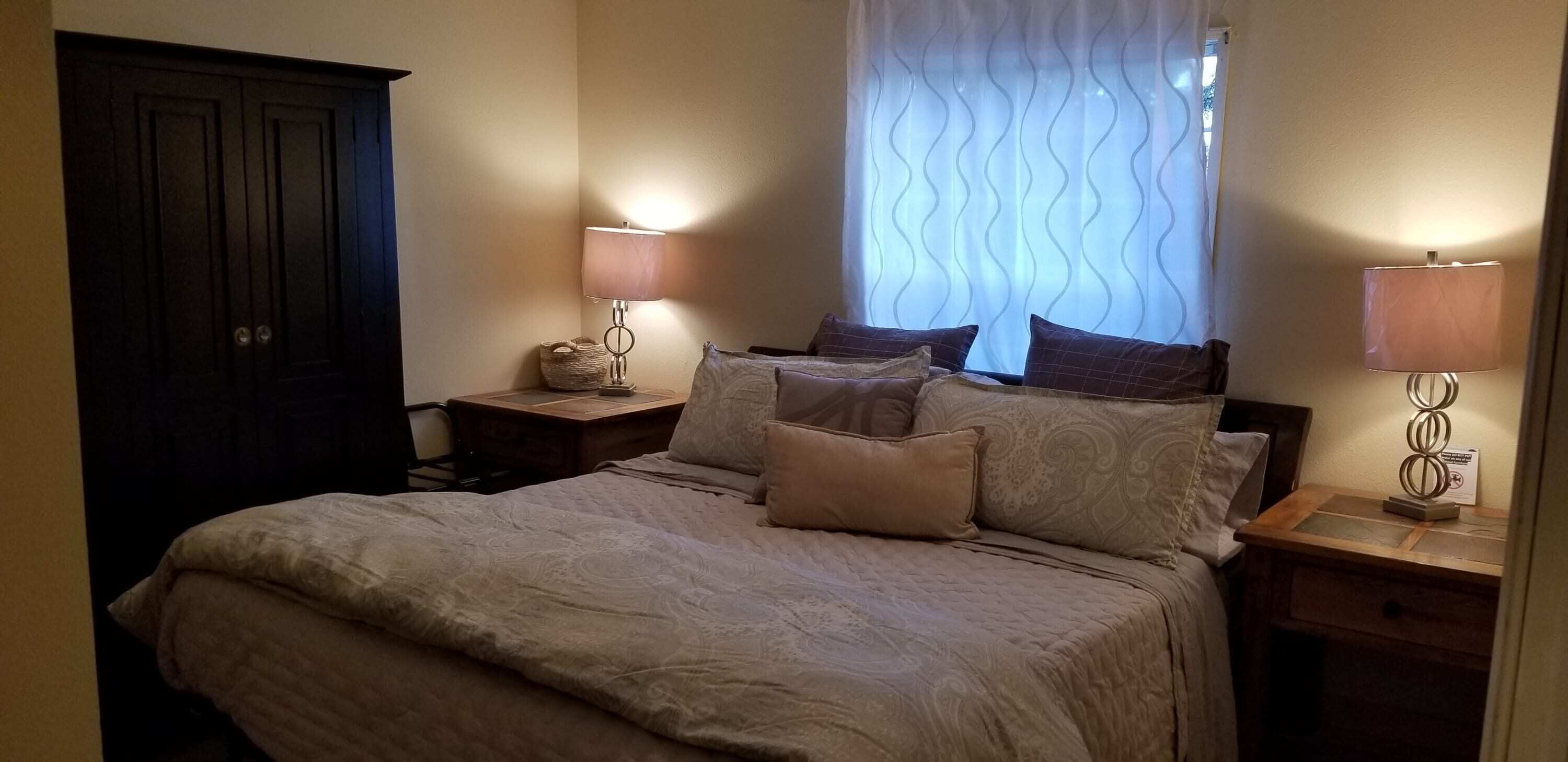 Un dormitorio con una cama grande, dos mesitas de noche, una mesilla y una ventana con cortinas, además de una puerta.