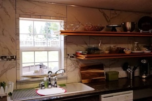 A kitchen with a window, marble backsplash, wooden shelves, and a sink with a dish rack.