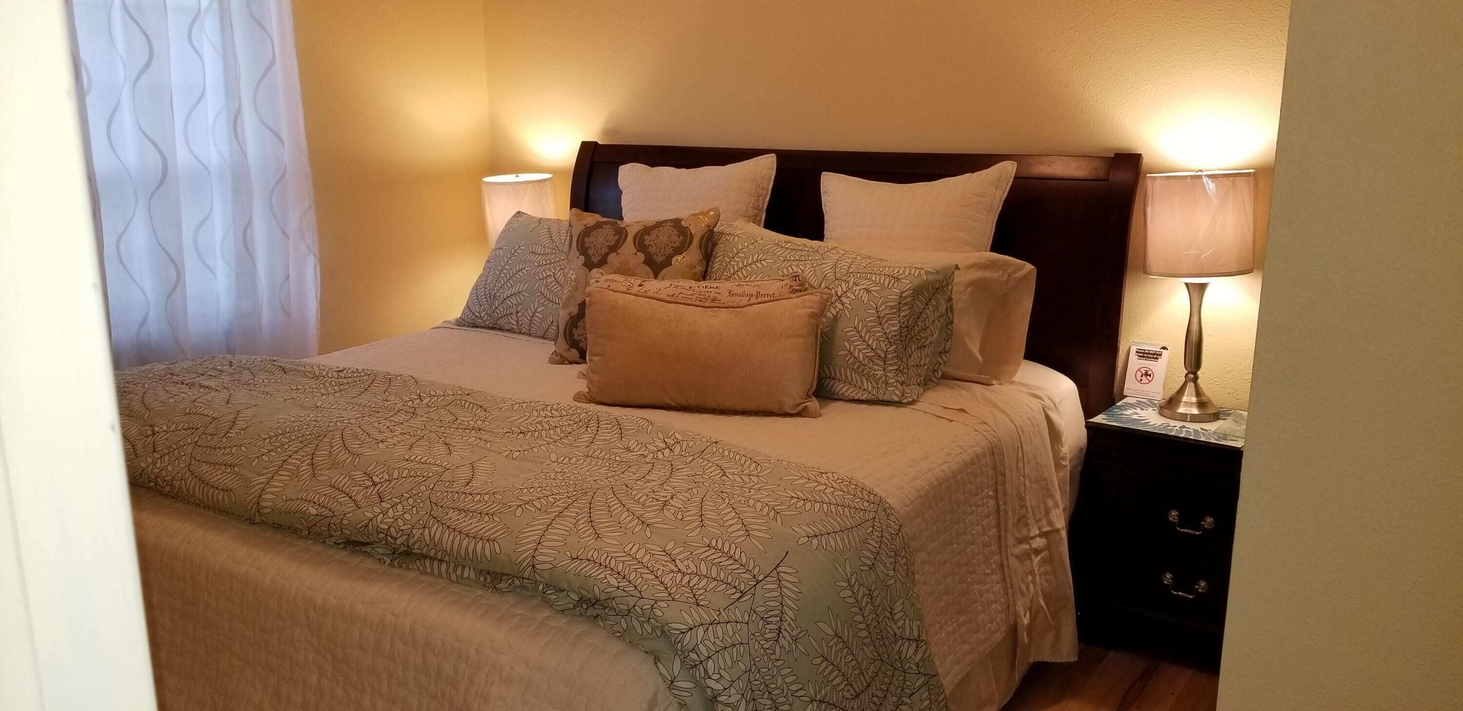 Una cama bien hecha con ropa de cama estampada y varias almohadas, una lámpara de noche y una mesita de noche con lámpara.