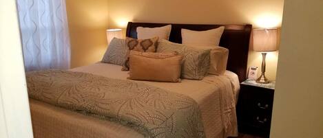 Una cama bien hecha con ropa de cama estampada y varias almohadas, una lámpara de noche y una mesita de noche con lámpara.