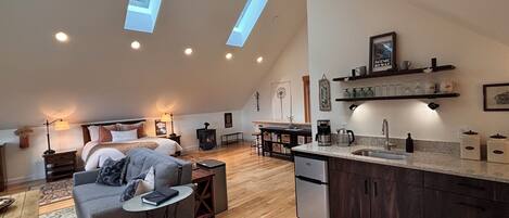 A loft-style bedroom with a bed, a gray sofa, a small table, and a heater.