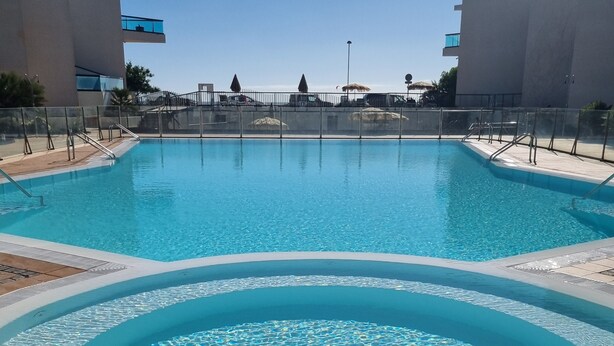 A clear blue swimming pool with a circular hot tub in the foreground, surrounded by a fenced area with a view of buildings in the background. - El Cotillo Sonando,Couples only, Romantic,Minutes from swim/surf,Well equipped. (El Cotillo)