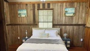 Une chambre avec un mur lambrissé en bois, un lit avec une literie blanche, une table de chevet avec une lampe et une fenêtre avec des stores.
