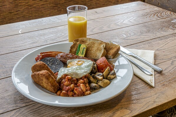 Un plato de comida para el desayuno que incluye tostadas, huevos, frijoles y salchichas, con un vaso de jugo de naranja sobre una mesa de madera.