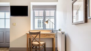 A room with a wooden desk, a chair, a TV mounted on the wall, a window with a view of a building, and a radiator. - Charlcombe Inn (Bath)
