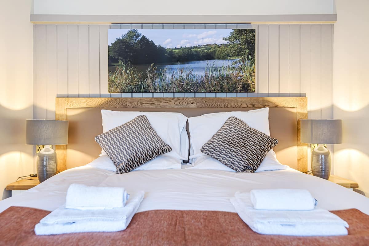 Habitación de hotel con cabecera de madera, dos almohadas estampadas, una cama con ropa blanca y vista a un lago a través de una ventana.
